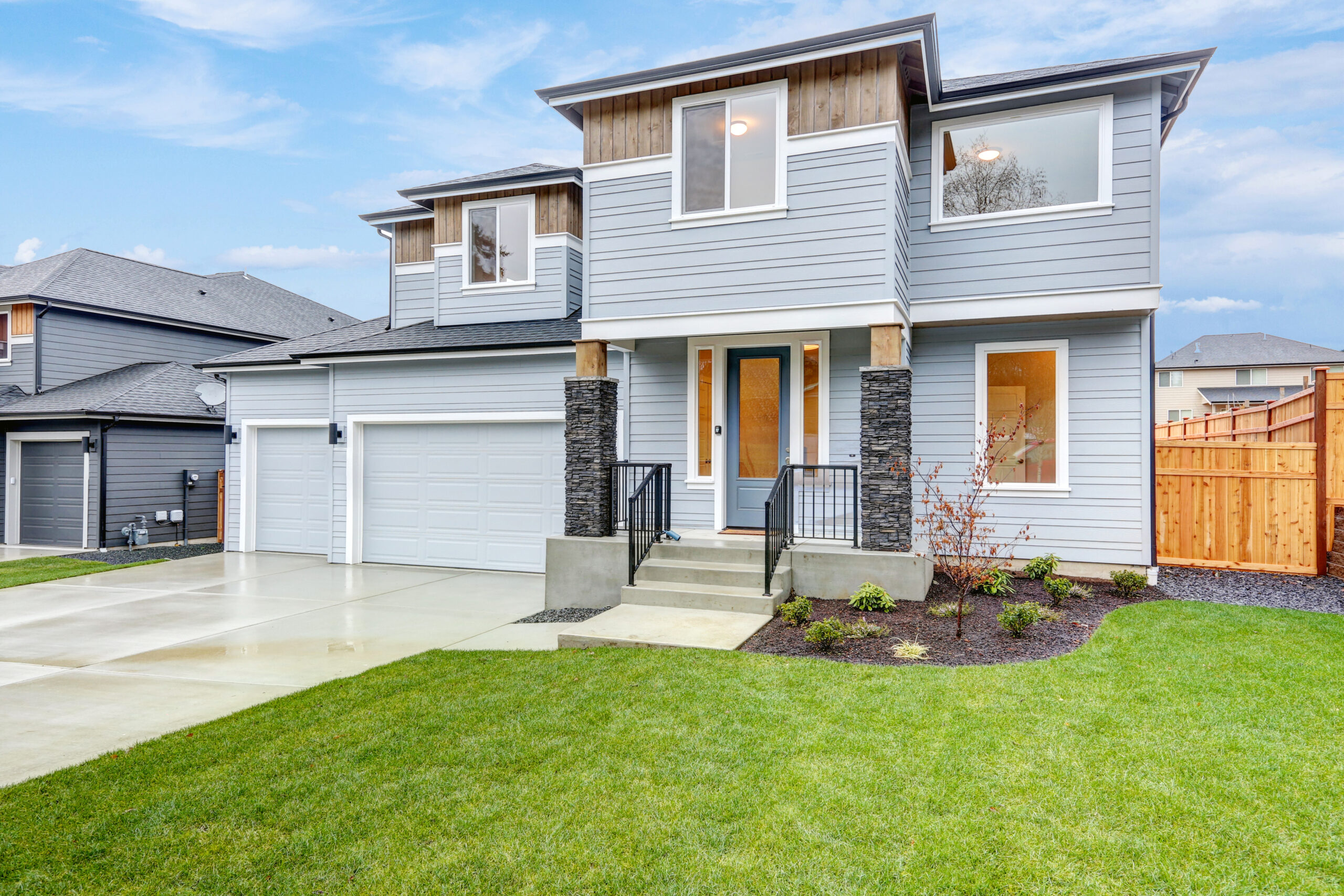 Two-story modern house with light gray siding, large windows, and a well-kept lawn. It features a two-car garage, stone accents, and a welcoming entrance.