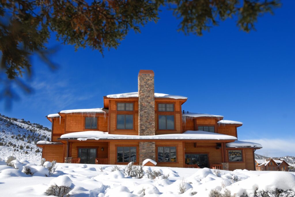 A cozy wooden cabin with a stone chimney is surrounded by snow under a clear blue sky. Snow-covered hills are in the background, with evergreen branches framing the scene.